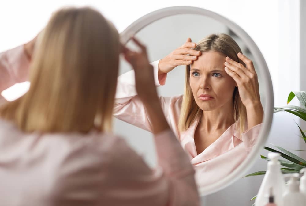 A woman examining her face in a mirror while recovering from a facelift procedure, looking for signs of swelling or asymmetry.