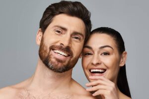 A smiling man with a beard and a woman with dark hair standing together, showcasing the distinct facial structures and hairlines that a surgeon must consider during a facelift procedure.