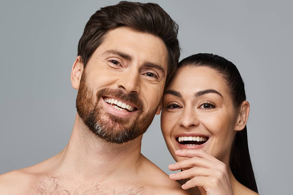 A smiling man with a beard and a woman with dark hair standing together, showcasing the distinct facial structures and hairlines that a surgeon must consider during a facelift procedure.