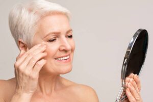 A woman with silver hair looking in a handheld mirror and gently touching the skin near her ear to check the healing progress of her facelift incisions.