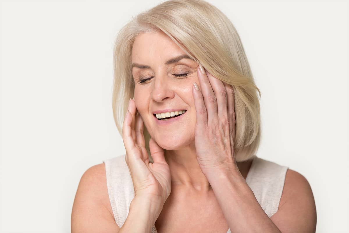 A woman with blonde hair gently cupping her face with both hands, illustrating the delicate touch required when applying topical treatments to facelift scars.