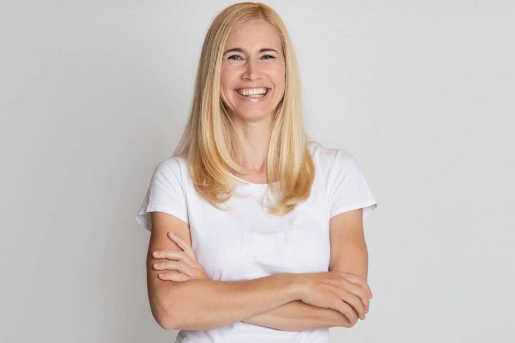 Smiling woman with smooth skin and crossed arms, representing long-term results of fat transfer versus dermal fillers.