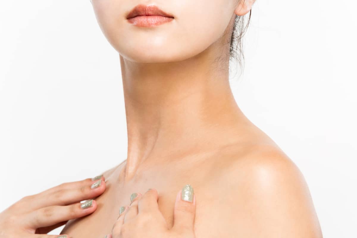 A close-up view of a woman's neck and jawline as she rests her hand with light green nails on her collarbone against a white background.