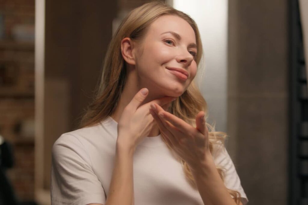 A smiling blonde woman in a white t-shirt gently touching her neck while looking into a mirror.