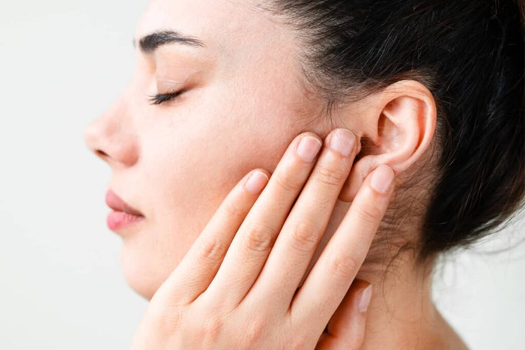A close-up side profile of a woman with dark hair and her eyes closed, as she gently lifts and supports the skin near her ear with her fingertips.